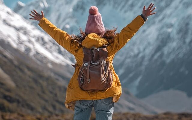 A woman in a yellow jacket and pink beanie, arms flung up toward snow-capped mountains, no phone in hand.